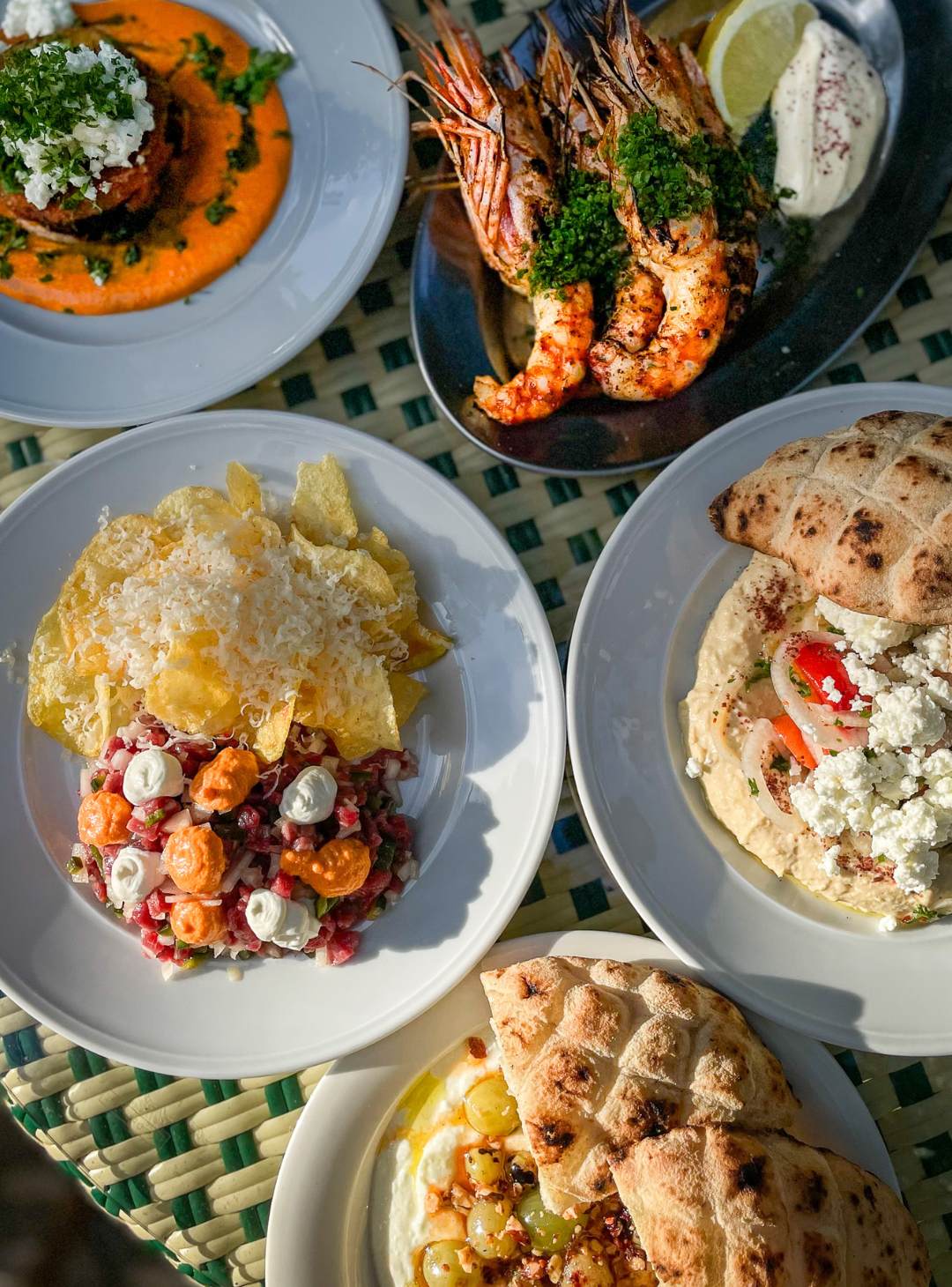 Uppdukat mezzebord i solljus med hummus och feta, grillade jätteräkor, råbiff och varmt bröd.
